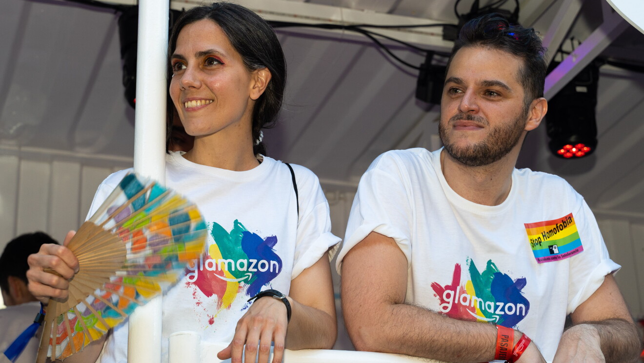 Eva y Simone en la carroza del desfile del Día internacional del orgullo LGTBI en Madrid. Los dos lucen la camiseta de glamazon y están apoyados en la barandilla de la carroza. Miran hacia su derecha con una sonrisa. De fondo se ve la carroza blanca y un foco de color rojo. 