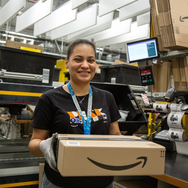 A young woman with brown hair and eyes is smiling, while holding a parcel in her hands working in a fulfillment center 