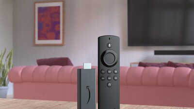 Em primeiro plano, uma Fire Stick TV aparece sobre uma mesa de madeira na sala de estar com o controle remoto e o USB para a TV. Ao fundo há uma TV pendurada na parede, um sofá rosa, um quadro pendurado na parede rosa e uma planta do lado esquerdo da sala