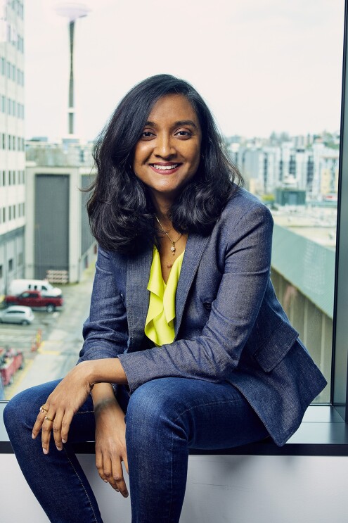 A woman sits on the ledge of window in an Amazon office building. She wears a blazer over a bright yellow top and jeans, smiling at the camera.