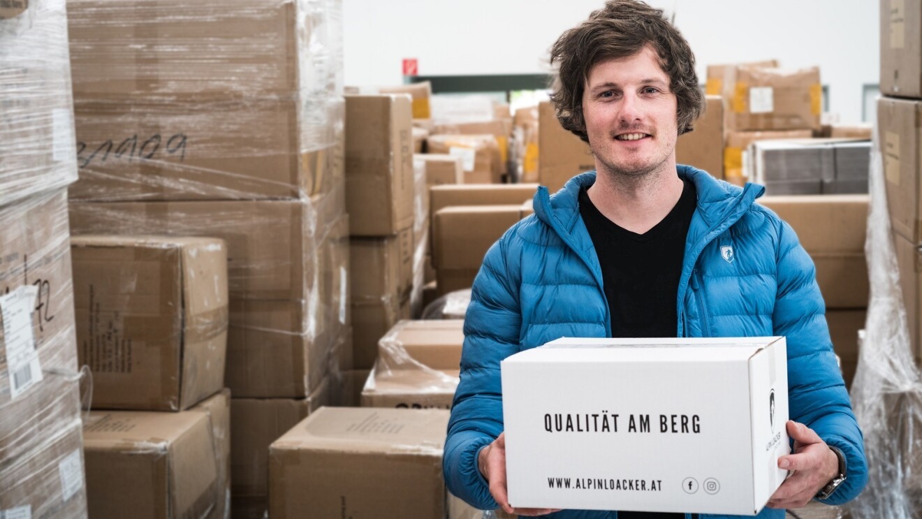 Ein Mann in einer blauen Jacke hält ein Packet von Alpin Loacker in den Händen, das die Aufschrift "Qualität am Berg" trägt. Im Hintergrund sind viele Kartons zu sehen.