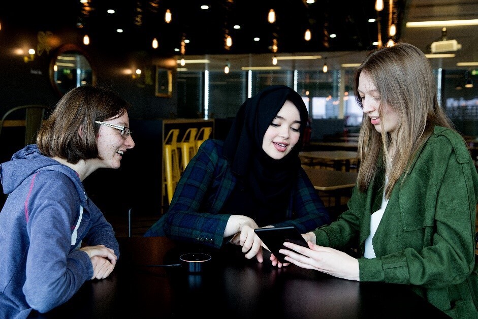 Three female students sat at a table smiling, talking and looking at a Kindle