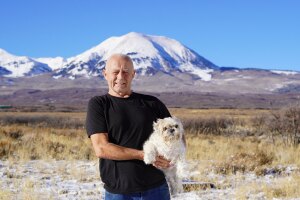 An image of a man smiling while holding a small white dog. There is a landscape behind him with a large, snow-capped mountain in the distance.