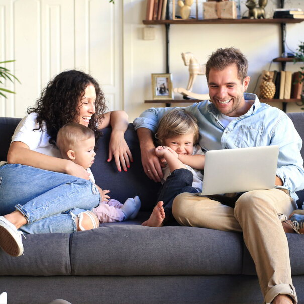 A family of four smiles and sits on a couch looking at a laptop.