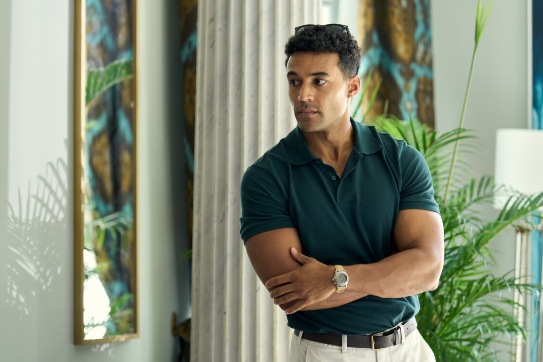 Man in green polo shirt standing with arms crossed in tropical-themed room