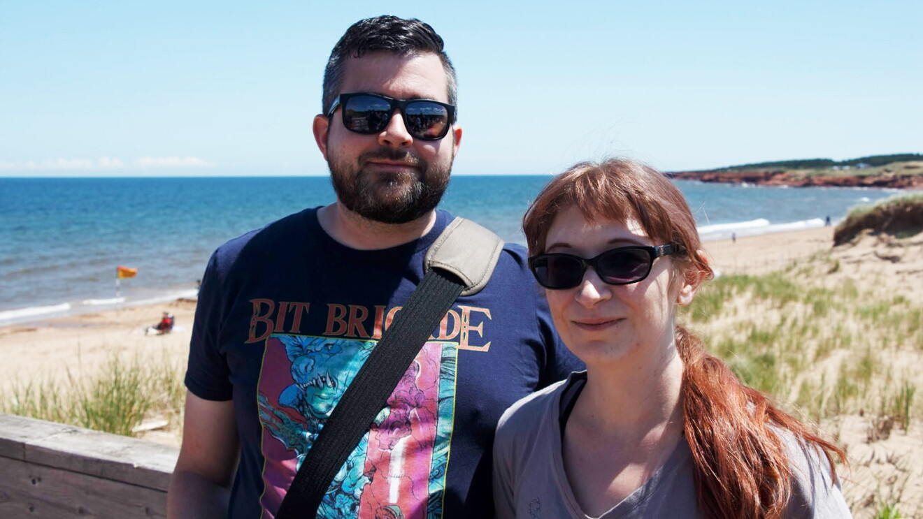 An image of a man and a woman smiling for a photo while standing on a platform in front of a beach.