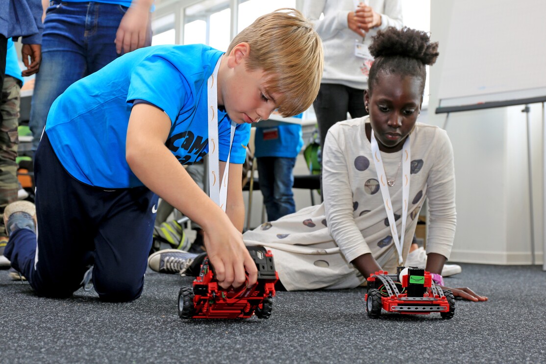 Ein blonder Junge und ein dunkelhäutiges Mädchen knien auf dem Boden und blicken auf zwei rote Modell-Transportroboter .