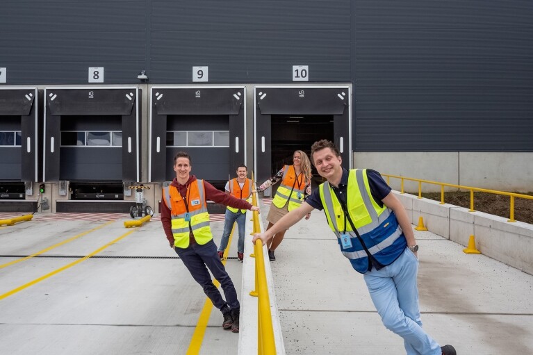 Vier Amazon Mitarbeiter:innen von Amazon Österreich in Warnwesten stehen an einem Geländer vor einem österreichischen Logistikzentrum von Amazon.