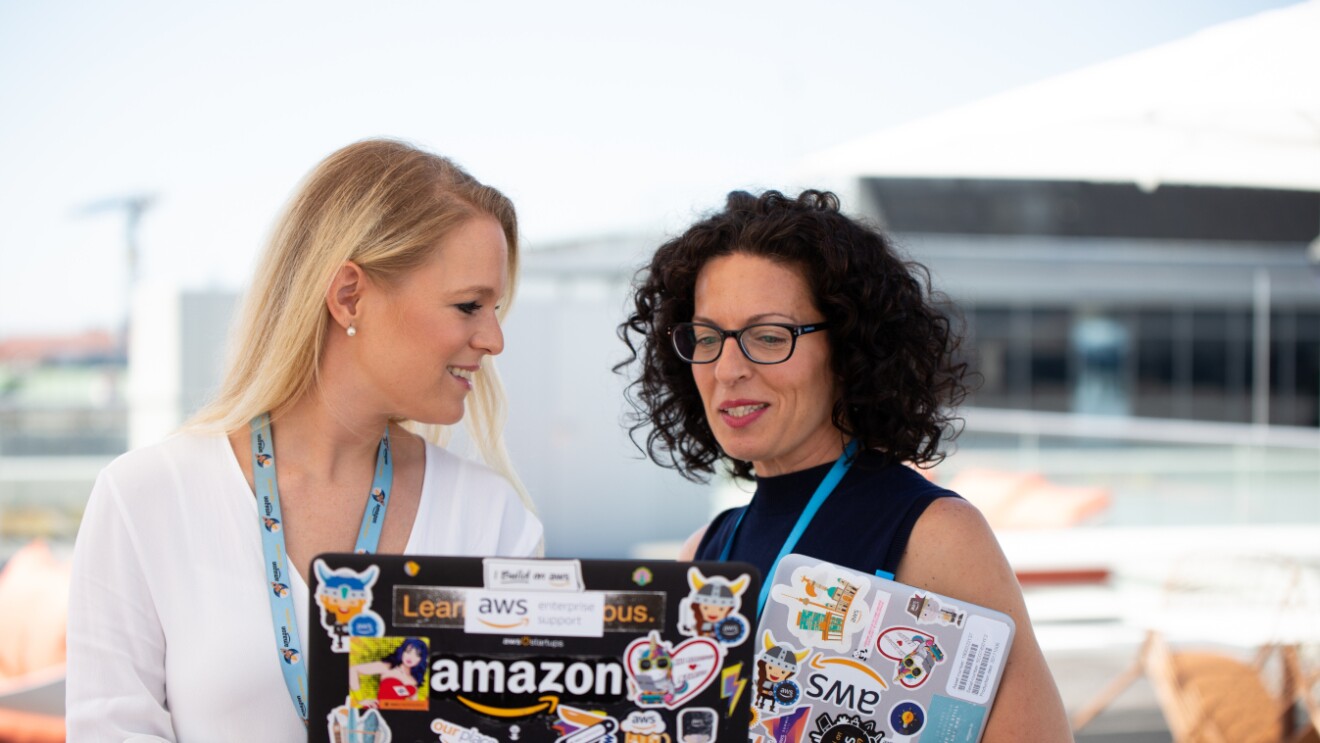 Zwei Frauen halten einen Laptop in den Händen auf dem Amazon steht. Sie sprechen miteinander.
