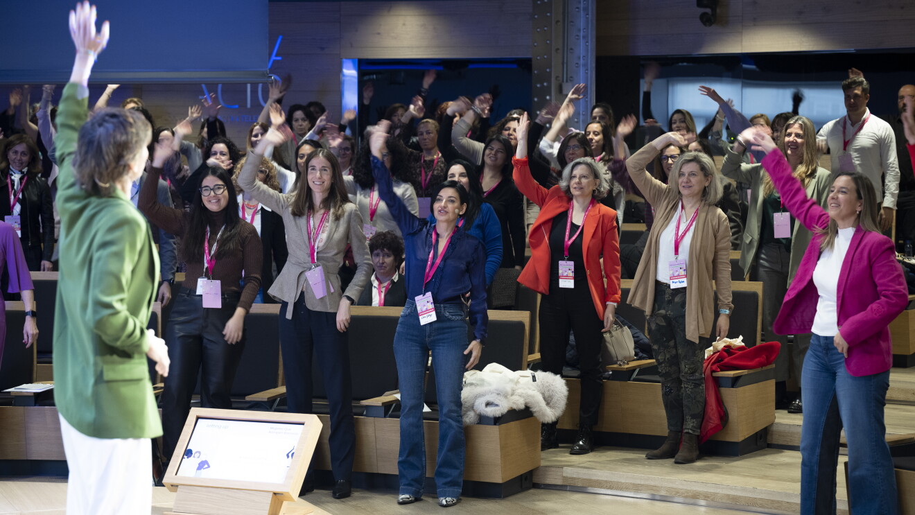 En un auditorio, aparece una mujer de espaldas con la mano izquierda levantada, Tiene el pelo corto y lleva una americana verde y un pantalón blanco. Delante de ella un auditorio lleno mayoritariamente de mujeres, se ve solo a dos hombres. Las mujeres están de pie con el brazo derecho levantado y todas llevan la acreditación con el colar de color rosa.