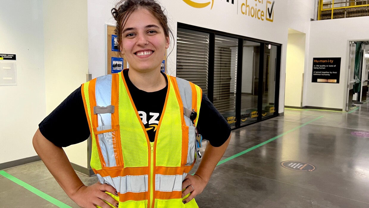 megan coghlan a process assistant at amazon standing in the foreground of an amazon fulfillment center. she is smiling with her hands on her hips and wearing yellow safety vest