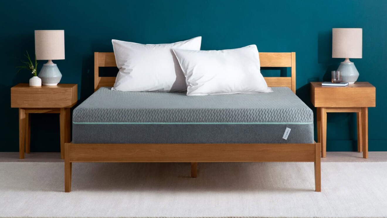 An image of a bedroom with a wood-frame bed and matching nightstand. The bed has a gray mattress on it with two white pillows.