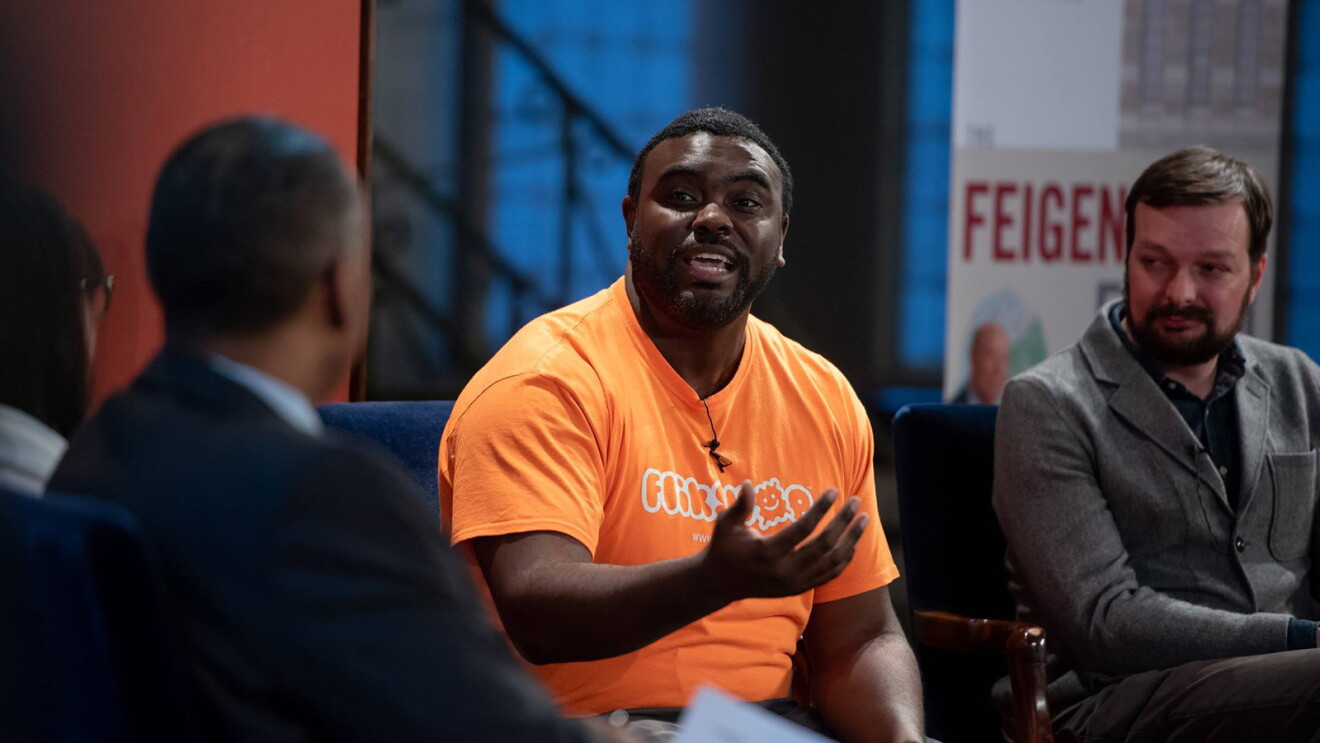 An image of a man speaking to a panel of people. He is wearing an orange shirt that says "Flikshop."
