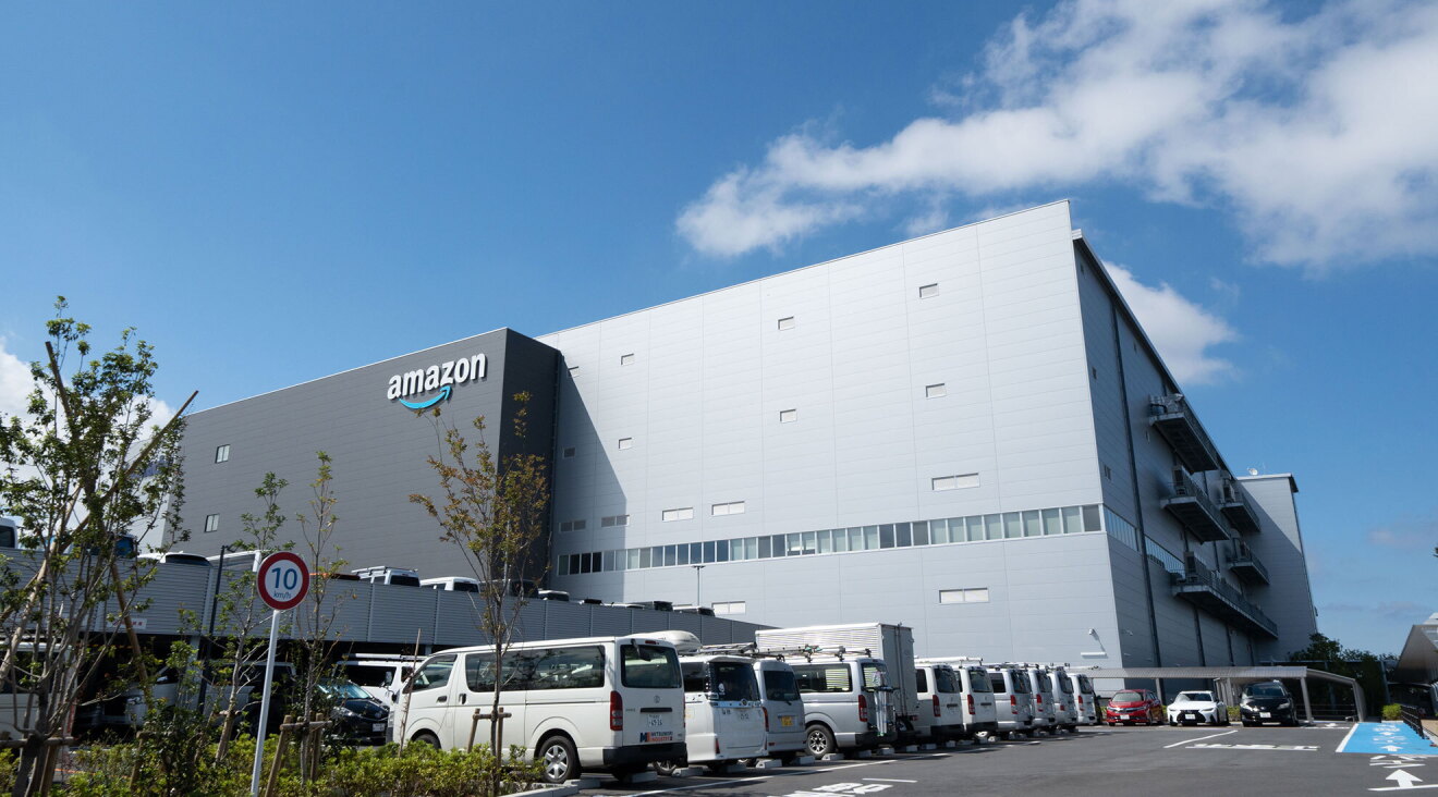 青い空をバックに建つ大きな建物。左側のダークグレーの壁にAmazonロゴ。右側の壁は薄いグレー。建物手間の立体駐車場に多数の車が停まっている。