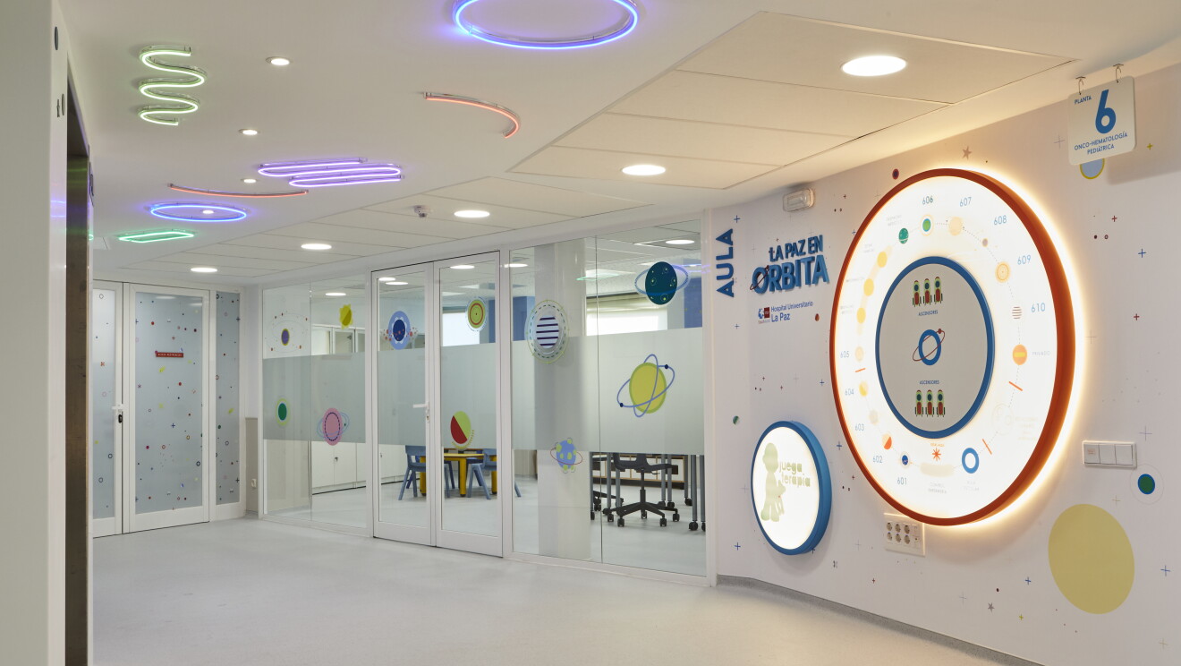 Aula formación de futuros astronautas del proyecto La Paz en Órbita del Hospital Universitario de La Paz. Desde el pasillo se ve una pared acristalada con vinilos que tienen dibujos de planetas y estrellas. El techo es blanco con luces de neon de colores dibujando formas geométricas. En la parte izquierda hay unas letras azules en mayúscula que pone Aula y La paz en Órbita. La pared tiene dibujos espaciales. 