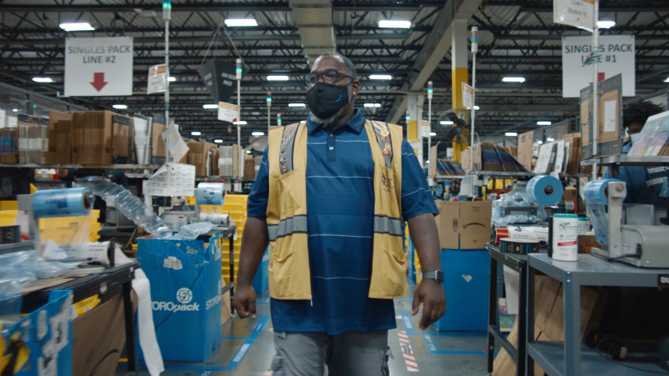 An image of a man walking through a walkway in an Amazon fulfillment center. He has a black mask on with the Amazon logo on it and a yellow safety vest and glasses. 