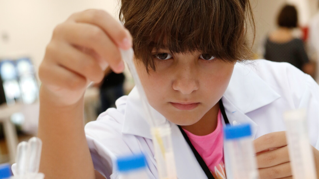Un chica de unos 11 años, con el pelo recogido y bata blanca, está colocando en una probeta un líquido. Tiene la mirada fija a la probeta y en la mano tiene una pipeta. Es un primer plano de la chica, El fondo difuminado. 