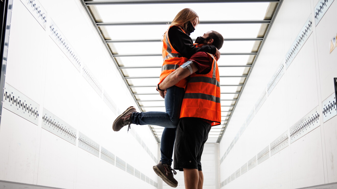 Sira Valeriano y Cristian Gamella. Foto de plano entero dentro de un túnel de carga de camiones de uncentro logístico. Los dos protagonistas van con una camiseta negra, él de manga corta y ella una sudadera y él va con el pelo corto y ella largo recogido con una coleta.Ël va con pantalón corto y ella con jeans. Cristian la está agarrando de los g´lúteos de tal forma que ella queda más alta. Ella le agarra del cuello y una de las pieras las tiene dobladas. 