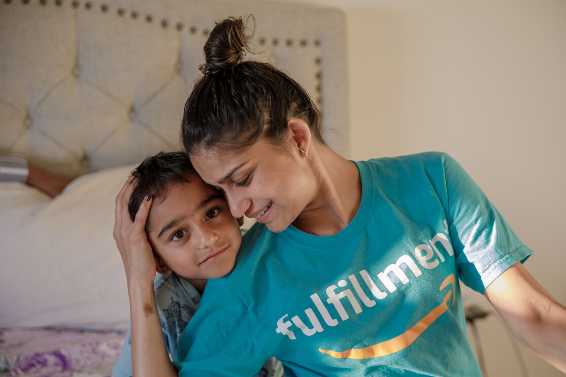 A young boy leans on the shoulder of a woman who cradles the side of his head with one hand. The woman has her hair up and wears a T-shirt with the Amazon smile logo and the word "fulfillment."