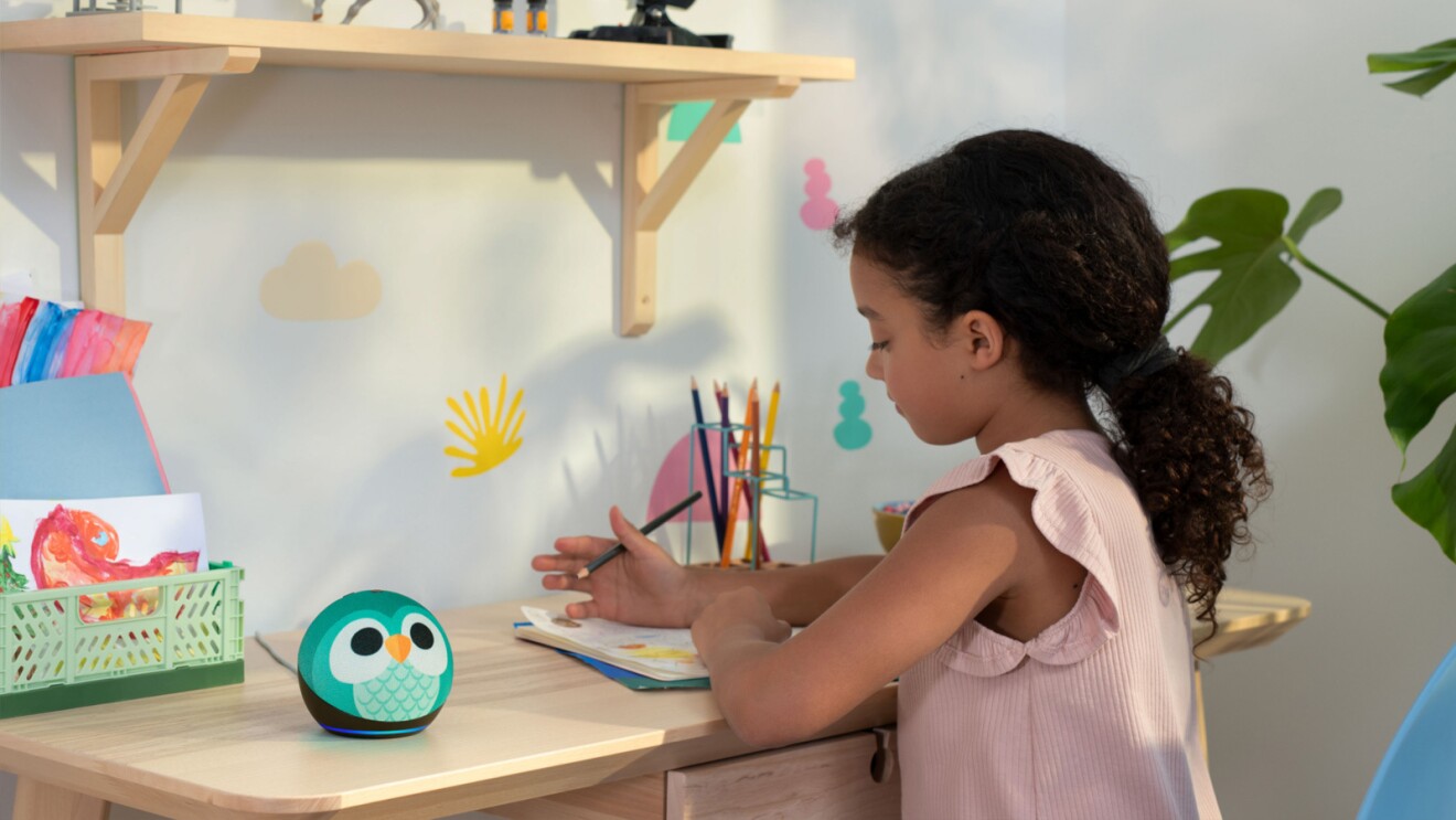 An image of a girl drawing in her desk with an Echo Dot Kids standing on the side of the desk. 