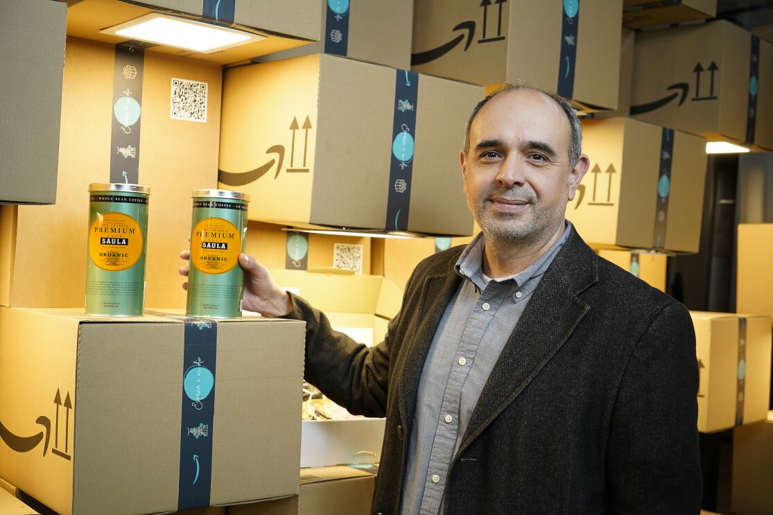En un escenario formado por cajas de Amazon de cartón. Octavi García, director de Marketing Digital de Café Saula, una pyme fundada por Lluís Saula en 1950 en el barrio del Raval de Barcelona y que es parte del nuevo escaparate de Amazon. Aparece delante de dos recipientes metálicos de Cafés Saula. Son de color verde con la etiqueta amarilla. Octavi agarra un recipiente con la mano derecha. Él lleva barba de dos días, una americana de pana, una camisa gris uy un jersey blanco debajo.