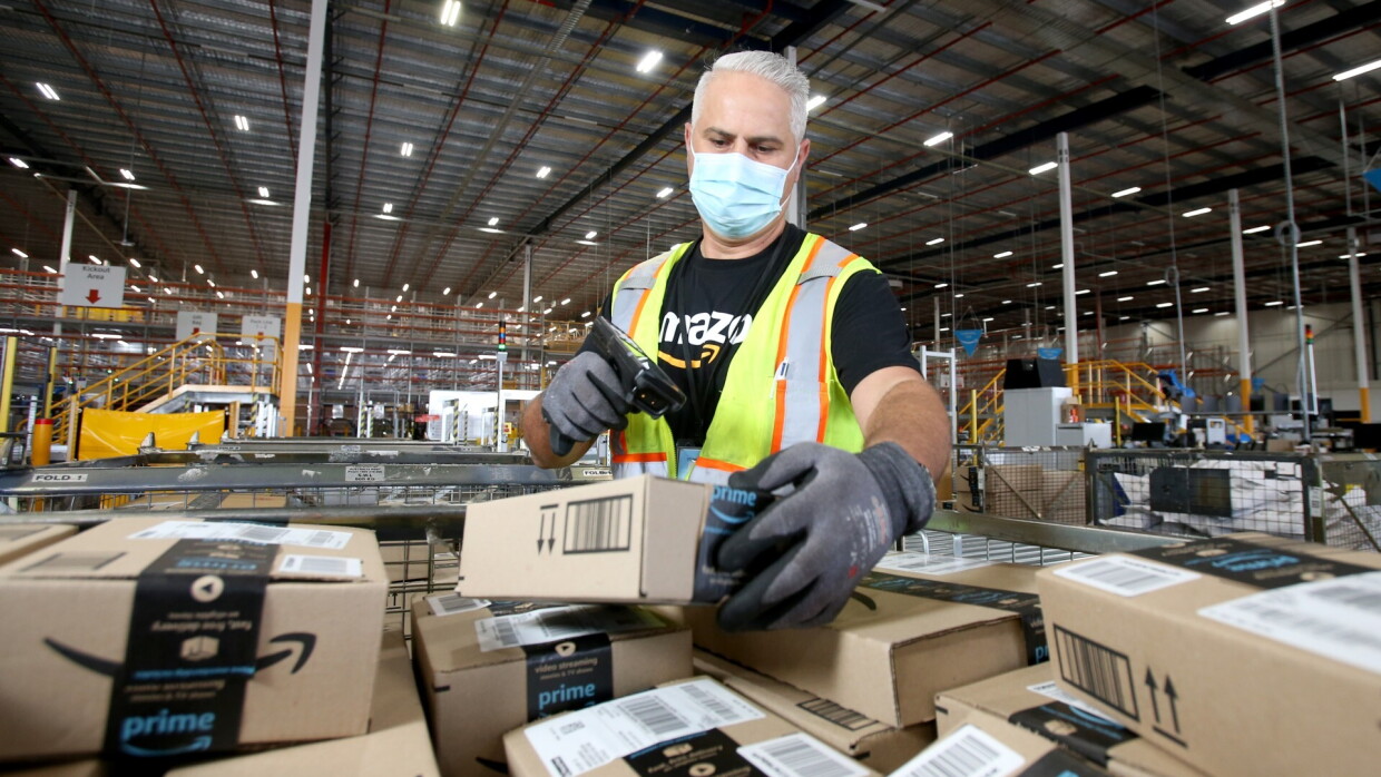 An Amazon employee scans packages in a fulfilment centre. He wears a black Amazon T-Shirt with a neon safety vest, and a face mask.