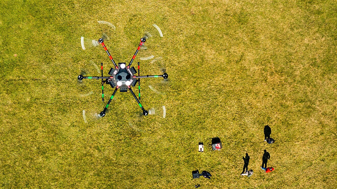 An image of a drone flying over a green field. People below are controlling it.