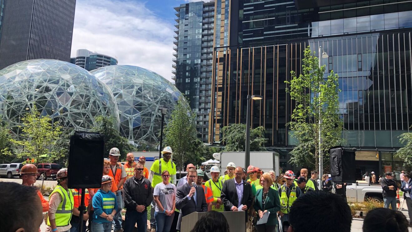 John Schoettler speaks into a microphone at a podium outside in front of a crowd. His team is behind him. They all stand in front of The Spheres.