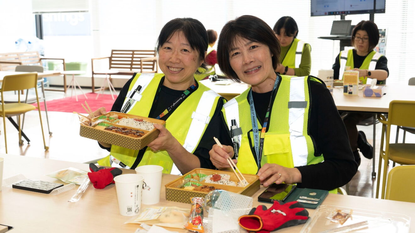 2人の女性がお弁当を見せている。彼女たちは作業用のベストを着ている。
