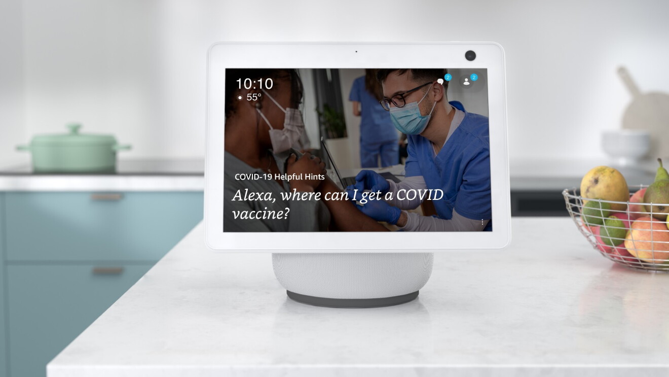 An Alexa device sits atop a white kitchen countertop and the screen reads "Alexa, where can I get a COVID-19 vaccine?" There is a wire basket filled with pears, limes, and apples next to the device. In the background there is a seafoam colored pot on the stove.