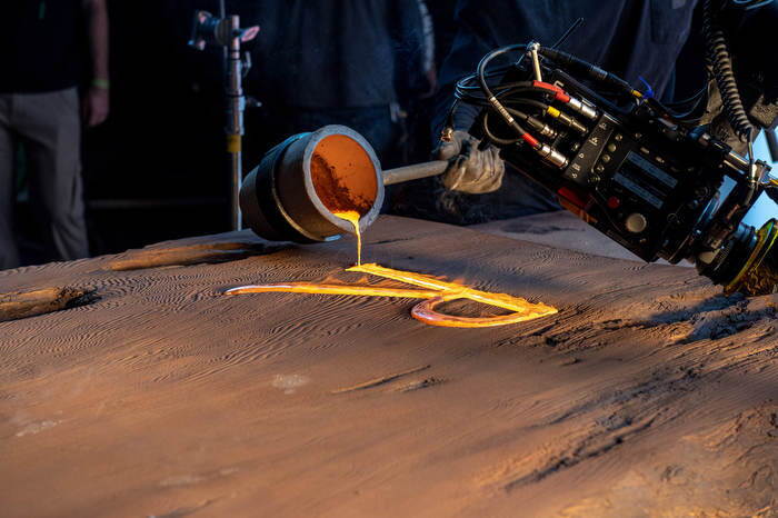 An image of a wood plank with am "R" carved into it. There is a welding tool pouring yellow, welded metal into the "R." This is a part of a larger sign that will eventually read "The Lord of the Rings" The Rings of Power."