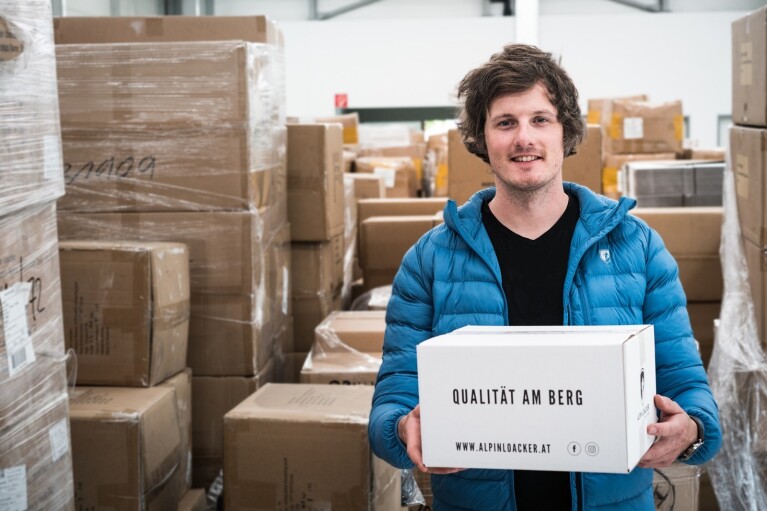 Ein Mann in einer blauen Jacke hält ein Packet von Alpin Loacker in den Händen, das die Aufschrift "Qualität am Berg" trägt. Im Hintergrund sind viele Kartons zu sehen.
