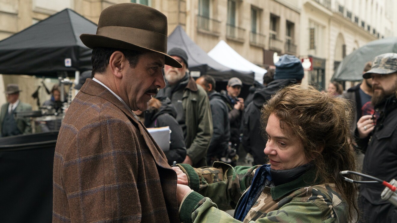An image of a woman fixing a part of an actor's costume. They are standing in the middle of a filming set in a city street.