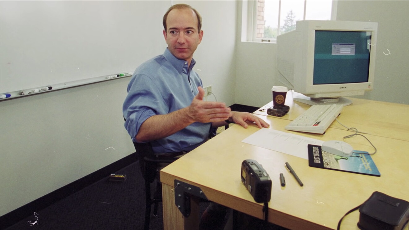 Jeff Bezos, muy joven, en su habitación de trabajo. De fondo una ventana y una pizarra blanca con los rotuladores de colores. Bezos está sentado en una silla negra y delante de una mesa de color madera. En la mesa hay un ordenador de mesa antiguo, un teclado y un ratón con su alfombrilla. En la mesa también un café con vaso de cartón, una cámara, un bolígrafo, un tapón de bolígrafo, una papel y una grabadora.