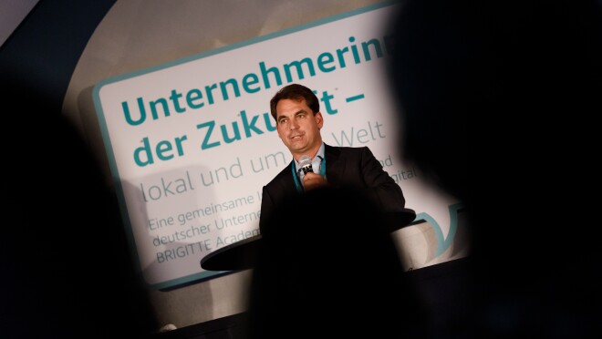 Nicholas Denissen steht auf der Bühne am Rednerpult. Er hält eine Laudation. Er ist aus dem Publikum heraus fotografiert worden. Im Vordergrund sind unscharfe Köpfe des Publikums zu erkennen.