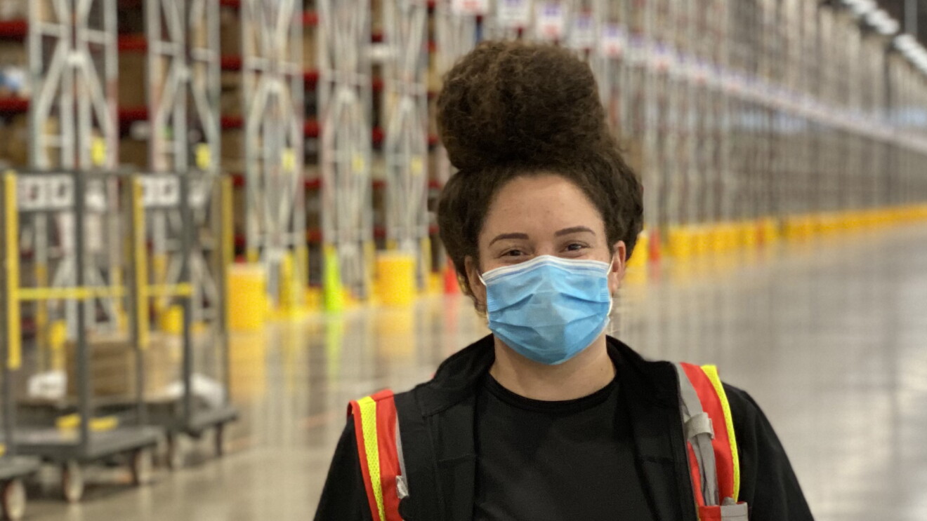 A woman wearing a safety vest and a mask stands in a warehouse space.