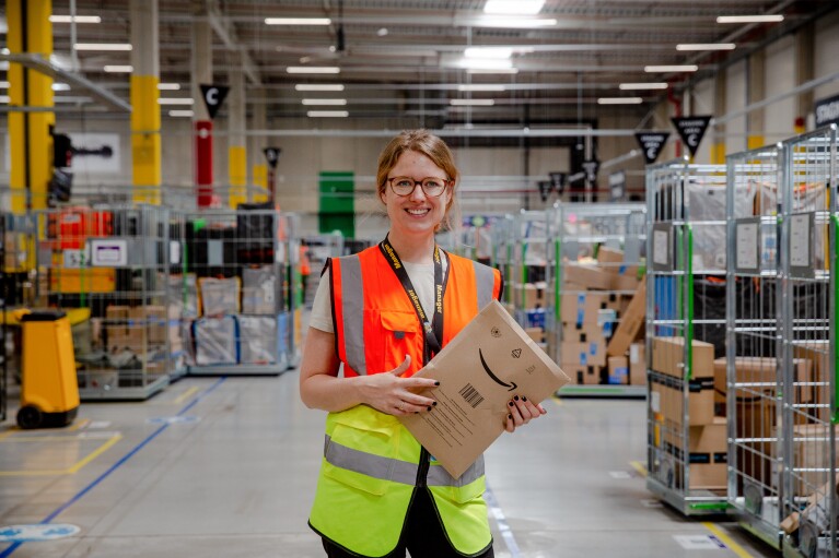 Regionalleiterin Miriam Enzi trägt eine gelb-orangene Weste und hält ein Amazon-Versandpäckchen in der Hand, während sie in einem Logistikzentrum von Amazon in Österreich steht.