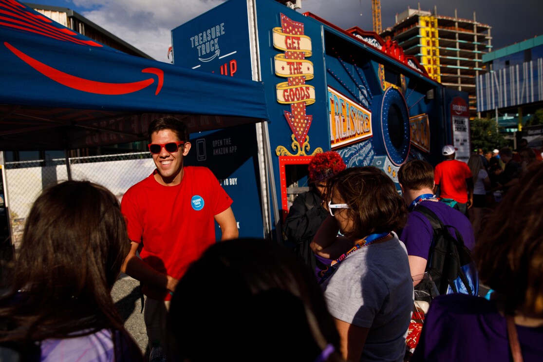 A man in a red tee shirt and sunglasses works at the Treasure Truck, handing out exclusive trading pins to athletes at the closing ceremony. The Treasure Truck is surrounded by people wearing Special Olympics clothing and lanyards.