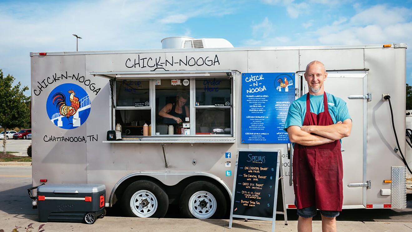 A man wearing a maroon apron stands with his arms crossed, in front of a silver CHICK-N-NOOGA food truck. A sign displays the specials in neon orange text on a black background.