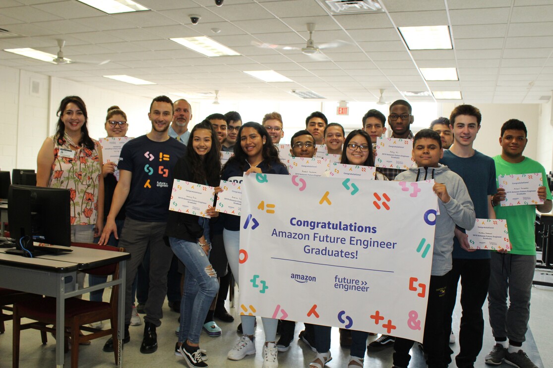 A group of students gathered in a classroom, standing behind an Amazon Future Engineer poster.