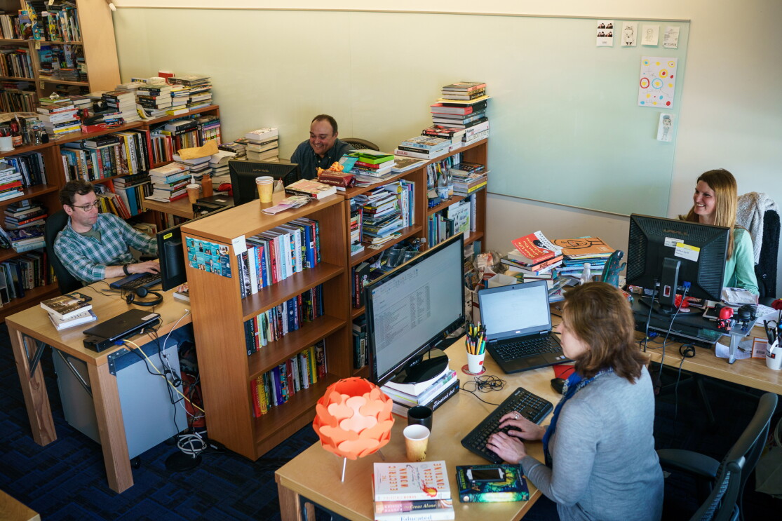 Two women and two men sit at desks and work at computers. They are in a shared office space with several full bookshelves, which also have small stacks of books on top of them.