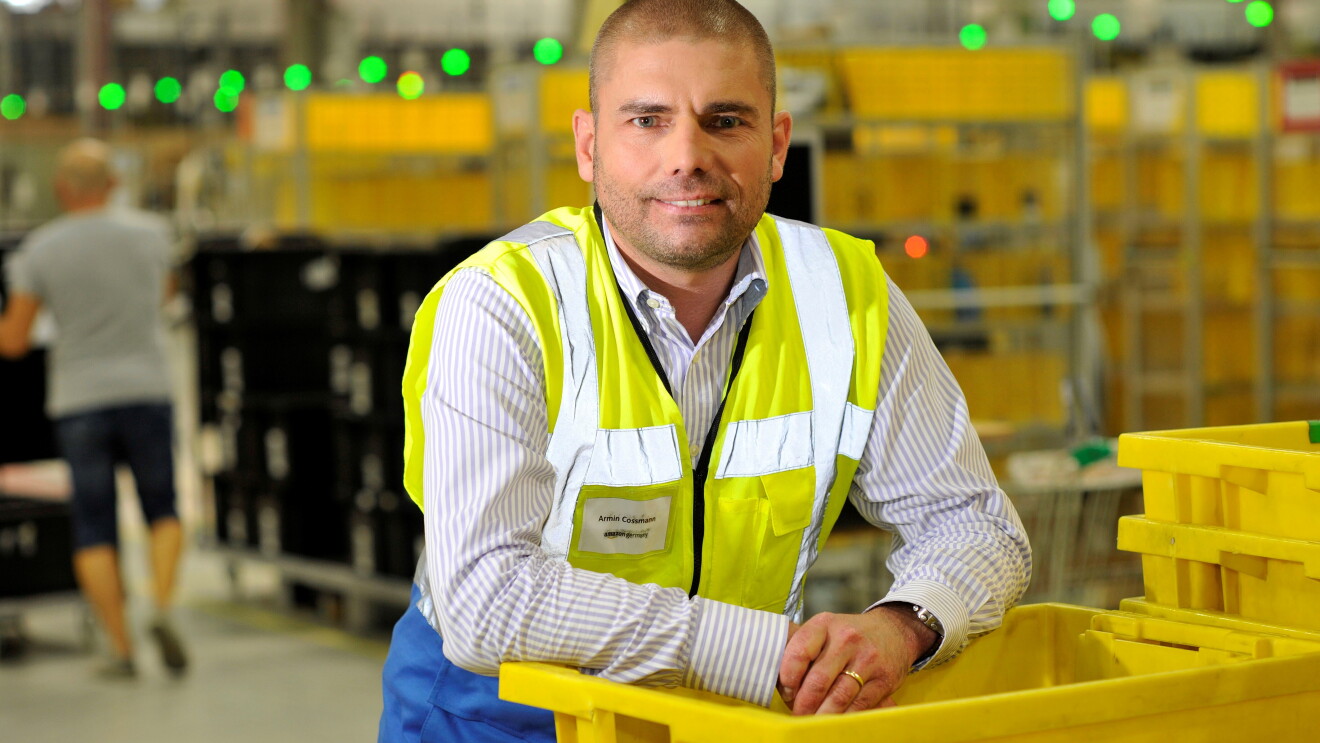 Ein Mann in gelber Sicherheitsweste lehnt sich lässig auf einem Stapler gelber Transportbehälter im Logistikzentrum