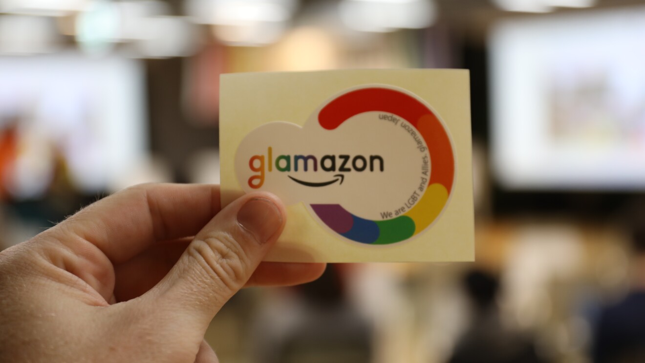 シールを3本の指で持つ左手のクローズアップ。背景はぼやけているが室内。シールは、レインボーカラーの輪と「glamazon」のロゴを組み合わせたもの。
