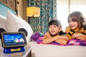 A picture of two kids laying on a hotel bed listening to an Echo smart speaker with Hey Disney! feature. 