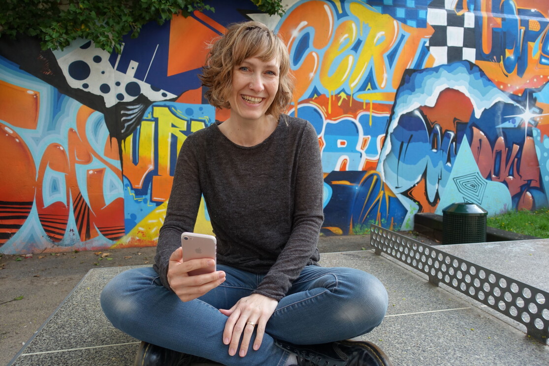 Eine Frau mit kurzen blonden Haaren und einem grauen Pullover sitzt auf einer Tischtennisplatte und lacht in die Kamera.