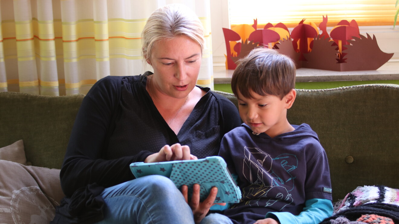 Eine blonde Frau sitzt mit einem Kinder auf einer Couch, beide schauen in ein Tablet.