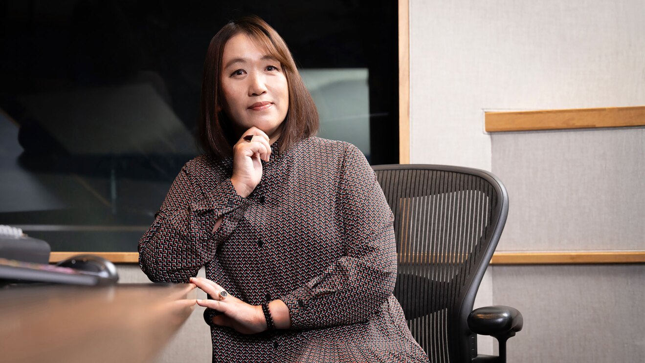 幾何学模様のワンピース姿で黒いオフィスチェアに座る女性。机に右肘をつき、右手を顎に軽く添えている。背景にはガラス越しにライトが消えた状態の録音ブースが見える。