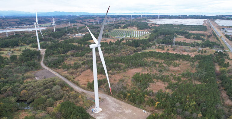 木々や畑が広がる緑の景色の中に、風力発電の白い風車が多数設置されている。風車以外にも道路や川などが見える。