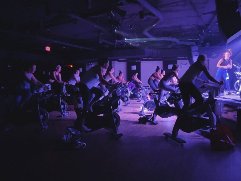 An image of a dark spin studio lit by a low, purple-tinted light. There are people riding stationary bikes and an instructor teaching at the front of the class.