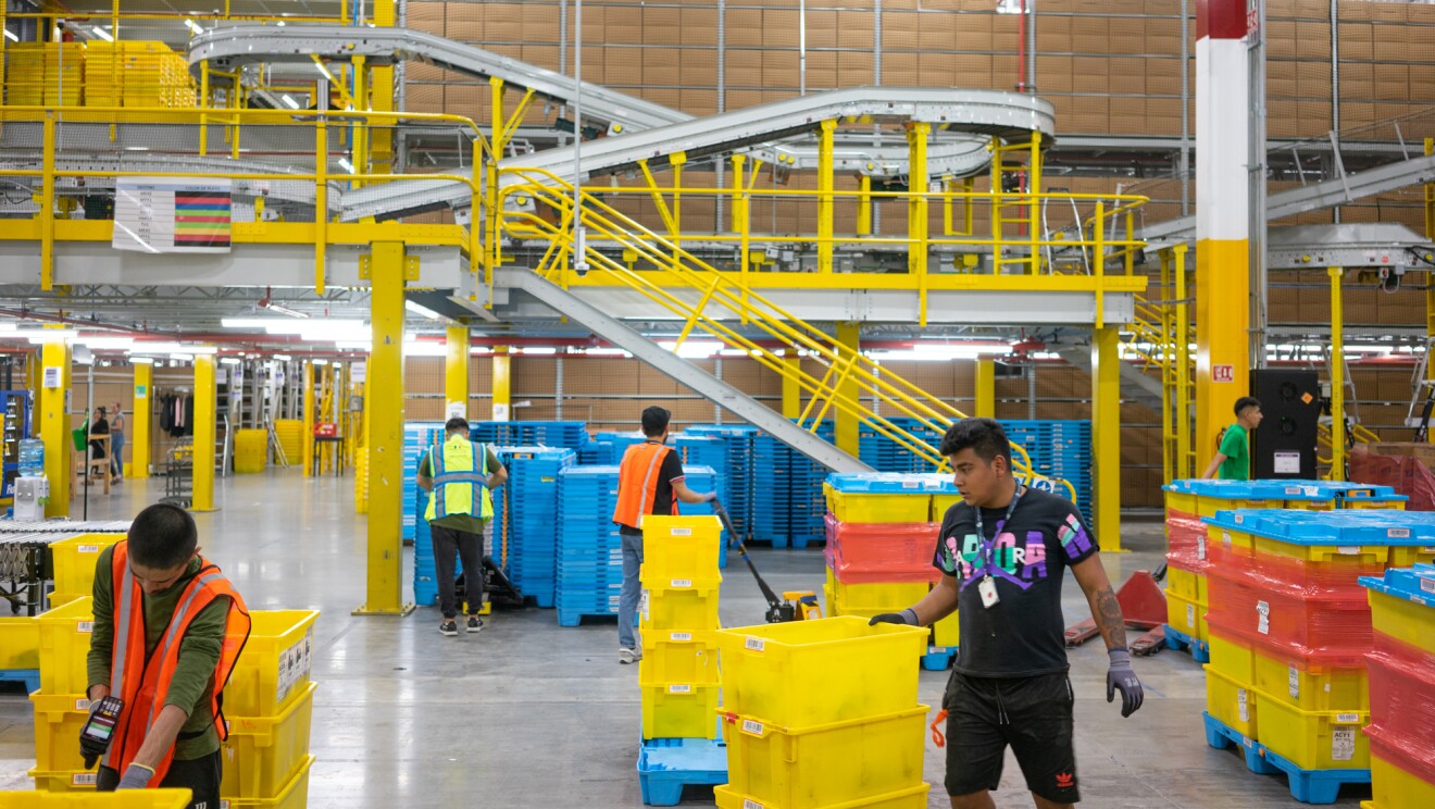 Imagen de un centro logístico de Amazon En primer plan se ven dos trabajadores manipulando cajas de plástico amarillas. De fondo tres personas más con toros semiautomáticos. Se ve de fondo una estantería y una cinta transportadora que termina en el suelo del centro logístico pero tienen un recorrido que llega a la parte superior de la foto, lo que podría ser el primer piso de la estantería.